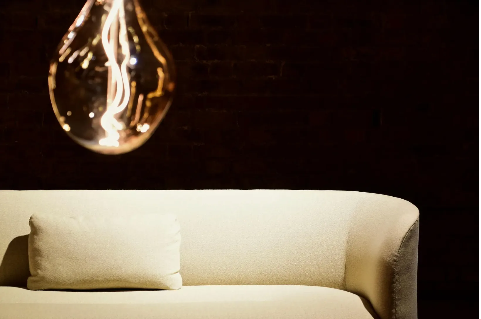 Beige curved Comfora sofa with a cushion, lit by an oversized glowing filament bulb against a dark brick wall.